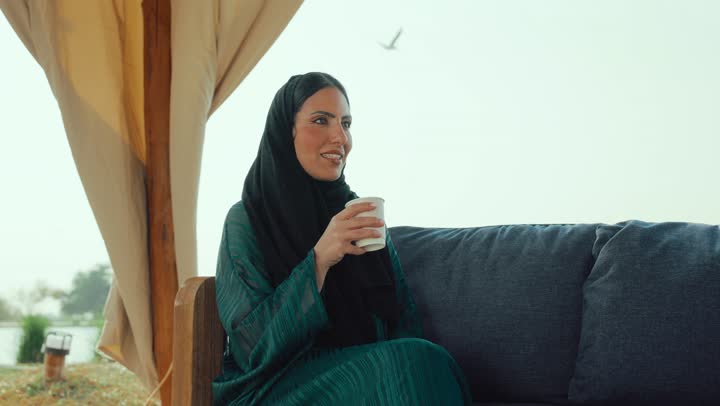 تصوير لامراة عربية خليجية اماراتية محجبة ترتدي العباءة تجلس على الأريكة في إحدى المقاهي الراقية تحمل كوب من القهوه، احتساء المشروب المفضل، سيده سعودية تستمع بلحظات الهدوء والراحة ، قضاء أوقات ممتعة ومسلية خارج المنزل, الشعور بالانتعاش والفرح