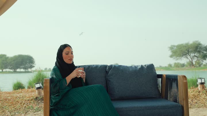 احتساء المشروب المفضل، سيده سعودية تستمع بلحظات الهدوء والراحة ، قضاء أوقات ممتعة ومسلية خارج المنزل, الشعور بالانتعاش والفرح، تصوير لامراة عربية خليجية اماراتية محجبة ترتدي العباءة تجلس على الأريكة في إحدى المقاهي الراقية تحمل كوب من القهوه