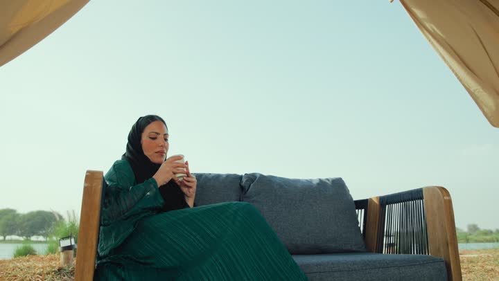 سيده سعودية تستمع بلحظات الهدوء والراحة ، قضاء أوقات ممتعة ومسلية خارج المنزل, الشعور بالانتعاش والفرح، احتساء المشروب المفضل، تصوير لامراة عربية خليجية اماراتية محجبة ترتدي العباءة تجلس على الأريكة في إحدى المقاهي الراقية تحمل كوب من القهوه