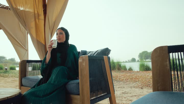 الشعور بالانتعاش والفرح، قضاء أوقات ممتعة ومسلية خارج المنزل, احتساء المشروب المفضل، تصوير لامراة عربية خليجية اماراتية محجبة ترتدي العباءة تجلس على الأريكة في إحدى المقاهي الراقية تحمل كوب من القهوه ، سيده سعودية تستمع بلحظات الهدوء والراحة
