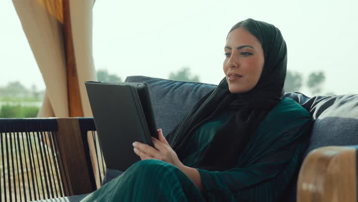 التواصل مع الأهل والأصدقاء، شابه سعودية تقوم باجراء مكالمه فيديو عبر التابلت، دمج التكنولوجيا الحديثة في الحياة اليومية، إلقاء التحية والترحيب والسلام، امراة عربيه خليجيه اماراتية مبتسمه ترتدي العباءه السوداء تجلس على الأريكة في إحدى المقاهي الراقية .