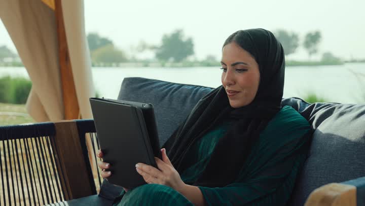 شابه سعودية تقوم باجراء مكالمه فيديو عبر التابلت، دمج التكنولوجيا الحديثة في الحياة اليومية، إلقاء التحية والترحيب والسلام، التواصل مع الأهل والأصدقاء، امراة عربيه خليجيه اماراتية مبتسمه ترتدي العباءه السوداء تجلس على الأريكة في إحدى المقاهي الراقية .