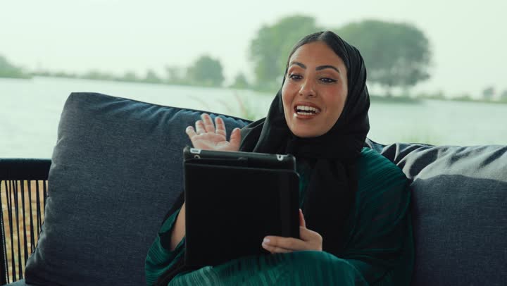 إلقاء التحية والترحيب والسلام، التواصل مع الأهل والأصدقاء، دمج التكنولوجيا الحديثة في الحياة اليومية، امراة عربيه خليجيه اماراتية مبتسمه ترتدي العباءه السوداء تجلس على الأريكة في إحدى المقاهي الرقية ، شابه سعودية تقوم باجراء مكالمه فيديو عبر التابلت