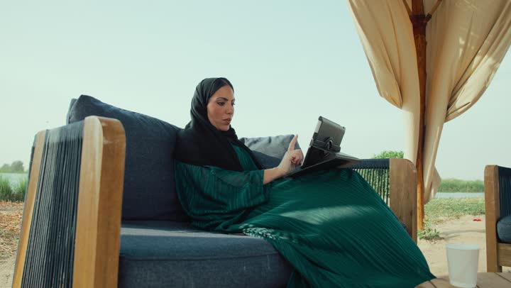 دمج التكنولوجيا الحديثة في الحياة اليومية، إلقاء التحية والترحيب والسلام، التواصل مع الأهل والأصدقاء، امراة عربيه خليجيه اماراتية مبتسمه ترتدي العباءه السوداء تجلس على الأريكة في إحدى المقاهي الرقية ، شابه سعودية تقوم باجراء مكالمه فيديو عبر التابلت