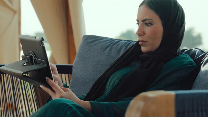 التواصل مع الأهل والأصدقاء، إلقاء التحية والترحيب والسلام، دمج التكنولوجيا الحديثة في الحياة اليومية، امراة عربيه خليجيه اماراتية مبتسمه ترتدي العباءه السوداء تجلس على الأريكة في إحدى المقاهي الرقية ، شابه سعودية تقوم باجراء مكالمه فيديو عبر التابلت