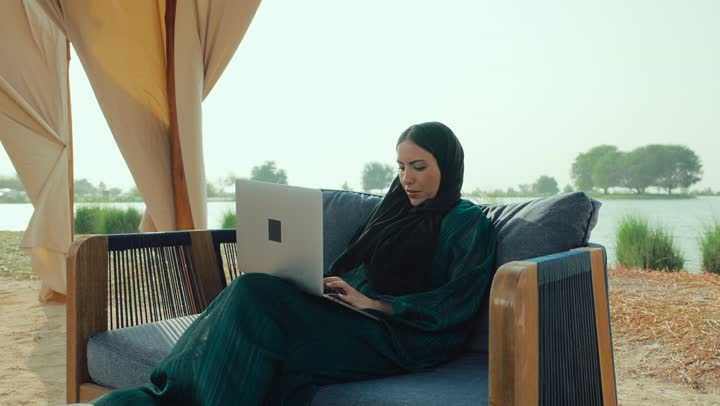 التسوق الالكتروني عبر الأونلاين، انجاز المهام عن بعد ، تصفح مواقع التواصل الاجتماعي، امراة عربيه خليجيه اماراتية مبتسمه ترتدي العباءه السوداء تجلس على الأريكة في إحدى المقاهي الراقية ، شابه سعودية تقوم تصفح الانترنت عبر جهاز الحاسوب المحمول .