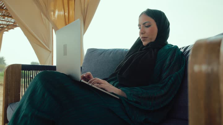 انجاز المهام عن بعد ، تصفح مواقع التواصل الاجتماعي، التسوق الالكتروني عبر الأونلاين، امراة عربيه خليجيه اماراتية مبتسمه ترتدي العباءه السوداء تجلس على الأريكة في إحدى المقاهي الراقية ، شابه سعودية تقوم تصفح الانترنت عبر جهاز الحاسوب المحمول .