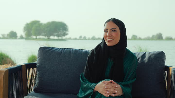 Hand gestures and different signs, greeting, peace and welcoming someone, a picture of an Arab Gulf Emirati woman wearing an abaya and hijab sitting on a sofa in a cafe in the Emirates, raising her hand in a welcoming gesture, gestures of happiness, joy and pleasure