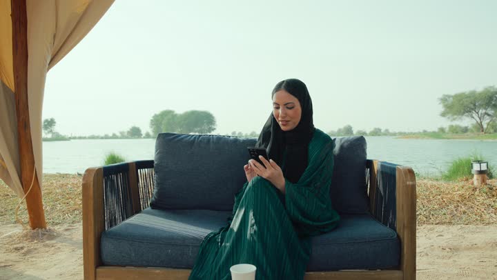 استعمال البرامج والتطبيقات الالكترونية، قضاء أوقات ممتعة ومسلية خارج المنزل، امراة عربية خليجية اماراتية ترتدي العباءة والحجاب تجلس على الأريكة في إحدى المقاهي الراقية تستمع بلحظات الهدوء والراحة ، سيده سعودية تقوم بتصفح الهاتف المحمول