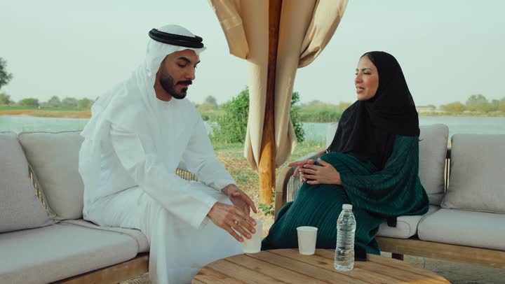 تبادل الاحاديث بين الأصدقاء، التسلية والترفيه في العطلة، الجلوس والاستمتاع بالمشروبات الساخنة والأعشاب الشتوية الدافئة ، ايماءات الاندماج فى الحديث ، تصوير لرجل عربي خليجي اماراتي يرتدي الكندورة والغترة البيضاء يجلس مع امراته  في المقهى ، ايماءات السعادة والسرور ، قضاء وقت ممتع فى الخارج  .