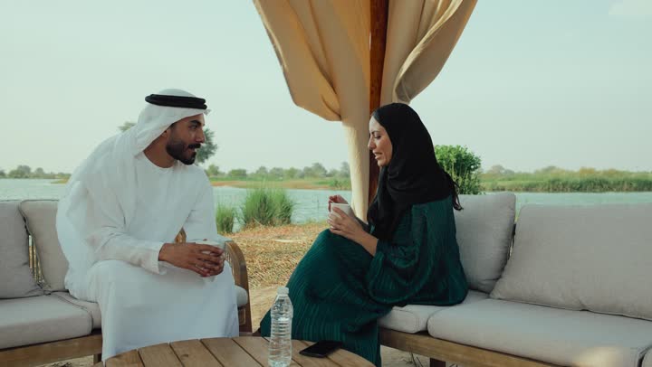 ايماءات السعادة والسرور ، قضاء وقت ممتع فى الخارج , الجلوس والاستمتاع بالمشروبات الساخنة والأعشاب الشتوية الدافئة ، ايماءات الاندماج فى الحديث ، تصوير لرجل عربي خليجي اماراتي يرتدي الكندورة والغترة البيضاء يجلس مع امراته  في المقهى ، التسلية والترفيه في العطلة، تبادل الاحاديث بين الأصدقاء .