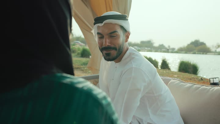 قضاء وقت ممتع فى الخارج , الجلوس والاستمتاع بالمشروبات الساخنة والأعشاب الشتوية الدافئة ، ايماءات الاندماج فى الحديث ، تصوير لرجل عربي خليجي اماراتي يرتدي الكندورة والغترة البيضاء يجلس مع امراته  في المقهى بايماءات السعادة والسرور ، التسلية والترفيه في العطلة، تبادل الاحاديث بين الأصدقاء .