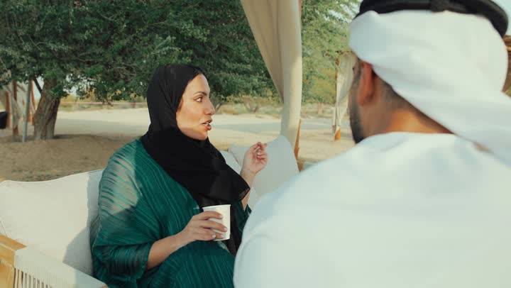 الجلوس والاستمتاع بالمشروبات الساخنة والأعشاب الشتوية الدافئة ، ايماءات الاندماج فى الحديث ، قضاء وقت ممتع فى الخارج ,  تصوير لرجل عربي خليجي اماراتي يرتدي الكندورة والغترة البيضاء يجلس مع امراته  في المقهى بايماءات السعادة والسرور ، التسلية والترفيه في العطلة، تبادل الاحاديث بين الأصدقاء .