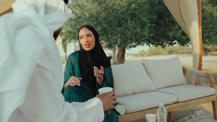 ايماءات الاندماج فى الحديث ، الجلوس والاستمتاع بالمشروبات الساخنة والأعشاب الشتوية الدافئة ، قضاء وقت ممتع فى الخارج ,  تصوير لرجل عربي خليجي اماراتي يرتدي الكندورة والغترة البيضاء يجلس مع امراته  في المقهى بايماءات السعادة والسرور ، التسلية والترفيه في العطلة، تبادل الاحاديث بين الأصدقاء .