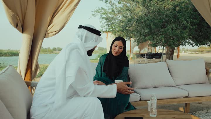 مشاركة المشروبات الساخنة والقهوة، قضاء وقت ممتع في الخارج، توثيق اللحظات السعيده ، تصوير لرجل عربي خليجي اماراتي يرتدي الكندورة والغترة البيضاء يجلس مع امراته  في المقهى بايماءات السعادة والسرور ،  التسلية والترفيه في العطلة، تبادل الاحاديث بين الأصدقاء، استخدام الهاتف المحمول .
