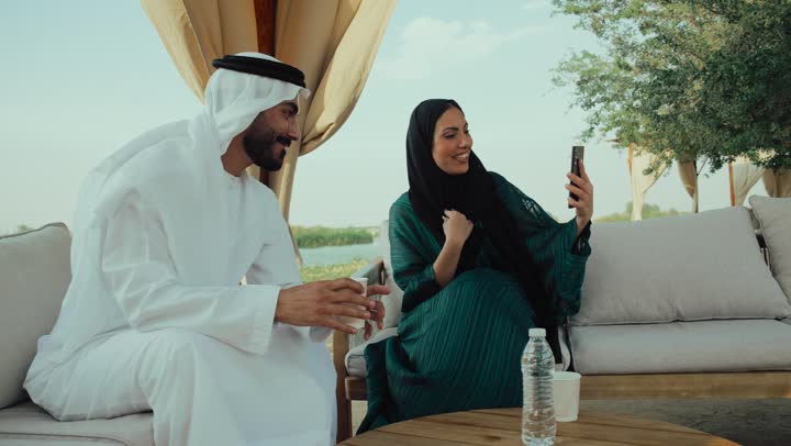 Having a good time outside, documenting happy moments, sharing hot drinks and coffee, a photo of an Emirati Gulf Arab man wearing a white kandura and ghutra sitting with his wife in a cafe with happy and pleased gestures, having fun and entertainment on vacation, chatting with friends, using a mobile phone.