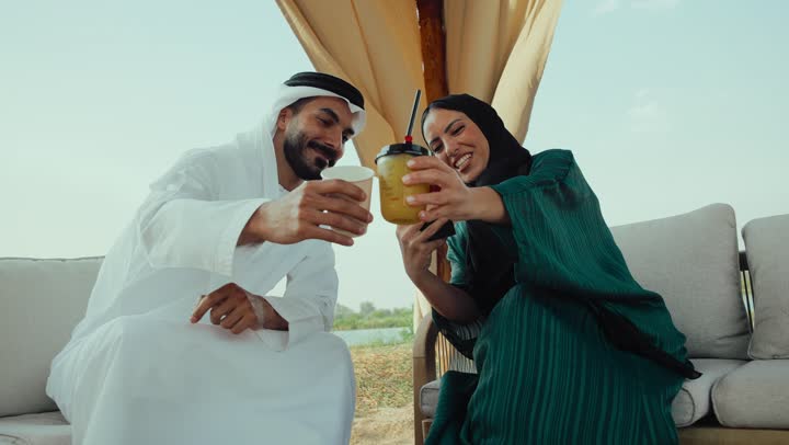 توثيق اللحظات السعيده ، مشاركة المشروبات الساخنة والقهوة، تصوير لرجل عربي خليجي اماراتي يرتدي الكندورة والغترة البيضاء يجلس مع امراته  في المقهى بايماءات السعادة والسرور ، قضاء وقت ممتع في الخارج، التسلية والترفيه في العطلة، تبادل الاحاديث بين الأصدقاء، استخدام الهاتف المحمول .