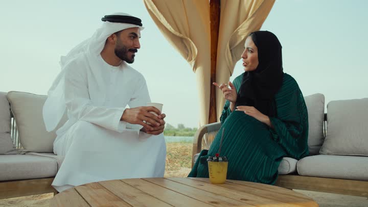 تصوير لرجل عربي خليجي اماراتي يرتدي الكندورة والغترة البيضاء يجلس مع امراته  في المقهى بايماءات السعادة والسرور ، قضاء وقت ممتع في الخارج، التسلية والترفيه في العطلة، تبادل الاحاديث بين الأصدقاء، مشاركة المشروبات الساخنة والقهوة، استخدام الهاتف المحمول .