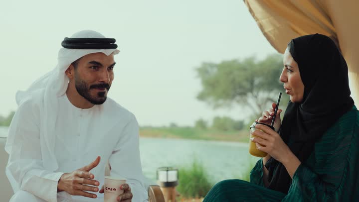 قضاء وقت ممتع في الخارج، التسلية والترفيه في العطلة، تبادل الاحاديث بين الأصدقاء، مشاركة المشروبات الساخنة والقهوة، استخدام الهاتف المحمول ، تصوير لرجل عربي خليجي اماراتي يرتدي الكندورة والغترة البيضاء يجلس مع امراته  في المقهى بايماءات السعادة والسرور .