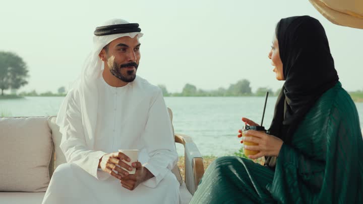 التسلية والترفيه في العطلة، تبادل الاحاديث بين الأصدقاء، مشاركة المشروبات الساخنة والقهوة، قضاء وقت ممتع في الخارج، استخدام الهاتف المحمول ، انجاز المهام عن بعد ، تصوير لرجل عربي خليجي اماراتي يرتدي الكندورة والغترة البيضاء يجلس مع امراته  في المقهى بايماءات السعادة والسرور .