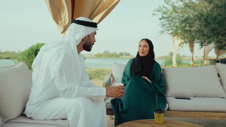 تبادل الاحاديث بين الأصدقاء، مشاركة المشروبات الساخنة والقهوة، قضاء وقت ممتع في الخارج، التسلية والترفيه في العطلة، استخدام الهاتف المحمول ، انجاز المهام عن بعد ، تصوير لرجل عربي خليجي اماراتي يرتدي الكندورة والغترة البيضاء يجلس مع امراته  في المقهى بايماءات السعادة والسرور .