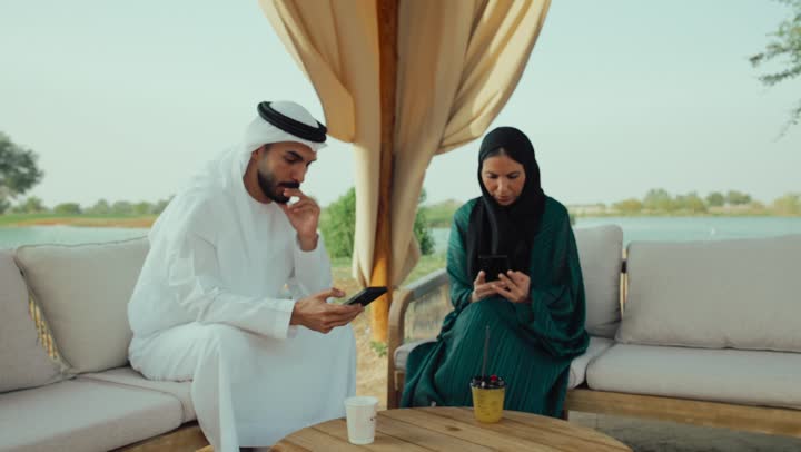 مشاركة المشروبات الساخنة والقهوة، تبادل الاحاديث بين الأصدقاء، قضاء وقت ممتع في الخارج، التسلية والترفيه في العطلة، استخدام الهاتف المحمول ، انجاز المهام عن بعد ، تصوير لرجل عربي خليجي اماراتي يرتدي الكندورة والغترة البيضاء يجلس مع امراته  في المقهى بايماءات السعادة والسرور .