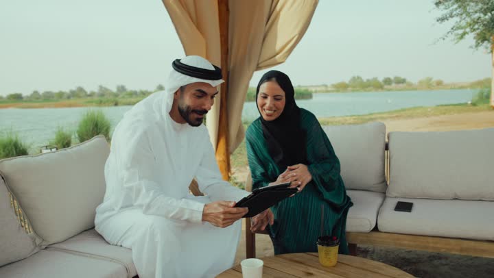 التواصل مع الاهل والاصدقاء عبر مكالمالت الفيديو ، استخدام التقنيات الحديثة ،تعابير السعادة والسرور بلمة الرفاق، التسامر والدردشة وقضاء وقت ممتع مع الأصدقاء، كوفي شوب راقي بالامارات، تصوير مقرب لرجل عربي خليجي اماراتي وامرأته يجلسون في المقهى .