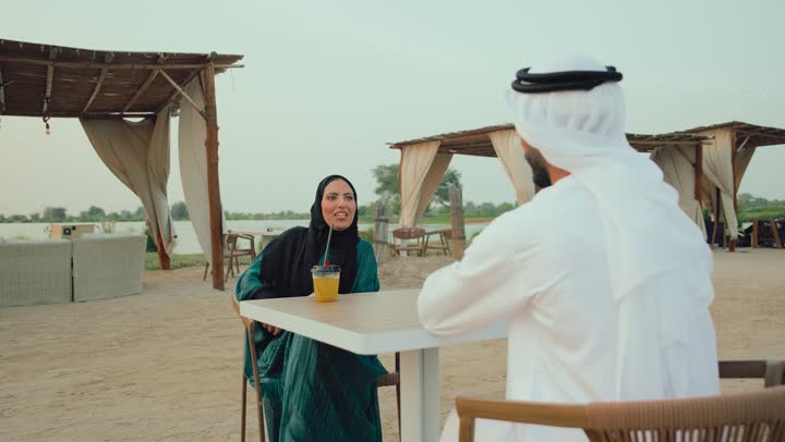 Close-up of an Emirati Gulf Arab man and his wife sitting in a cafe enjoying a conversation, gestures of pleasure and happiness, family dialogue and discussion, family entertainment activities and events, the concept of intimacy and love between family members, entertainment and recreation during holidays.