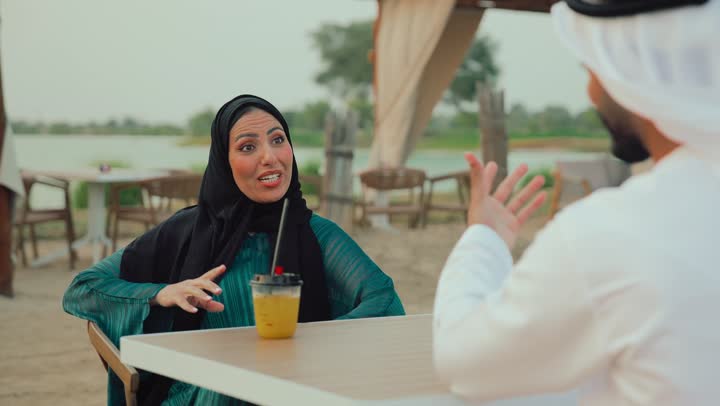 حوار ونقاش اسري، انشطة وفعاليات ترفيهية عائلية، مفهوم الألفة والمحبة بين أفراد الأسرة، التسلية والترفيه بالعطلات، ايماءات السرور والسعادة، تصوير مقرب لرجل عربي خليجي اماراتي وامرأته يجلسون في المقهى يستمتعون بالنقاش معاً .