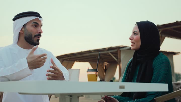 ايماءات السرور والسعادة، حوار ونقاش اسري، انشطة وفعاليات ترفيهية عائلية، مفهوم الألفة والمحبة بين أفراد الأسرة، التسلية والترفيه بالعطلات،  تصوير مقرب لرجل عربي خليجي اماراتي وامرأته يجلسون في المقهى يستمتعون بالنقاش معاً .
