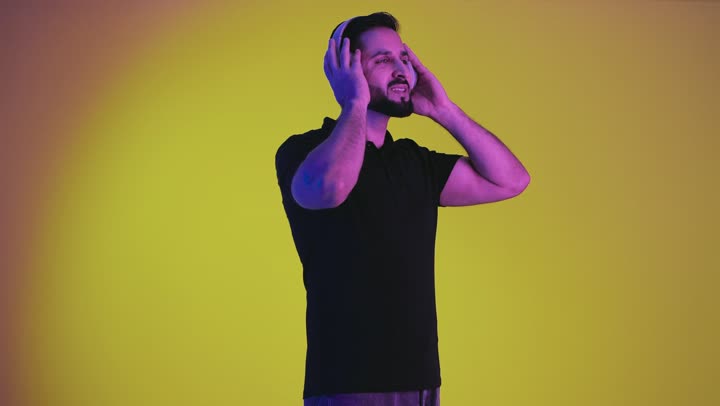 Artistic singing talents, an Arab Gulf Emirati man holding and holding his headphone, gestures and facial expressions indicating happiness and integration with the song, a portrait of a Saudi young man wearing casual clothes listening to music, a recording of a singing audio clip, a yellow background