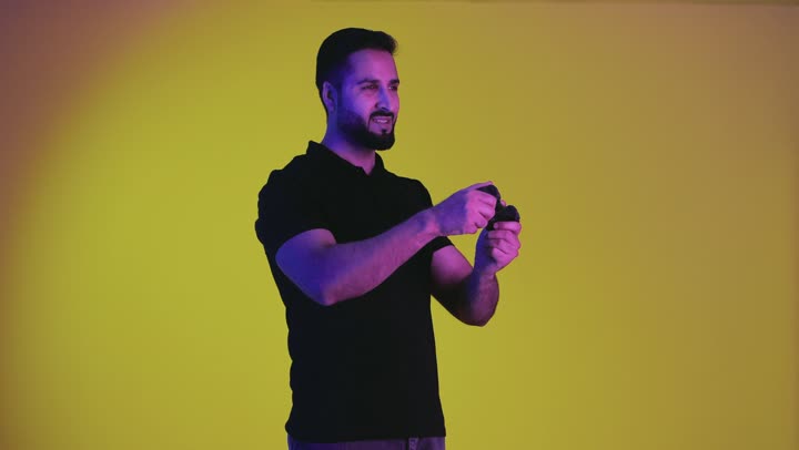 شاب عربي خليجي سعودي يمسك بيده عصا التحكم و يلعب ألعاب الفيديو بإيماءات و تعابير تدل على الفوز و السعادة ، بورتريه لرجل اماراتي يرتدي الزي الكاجوال يشعر بالمتعة و التسلية بألعاب الفيديو ، ألعب البلايستيشن الترفيهية المسلية ، خلفية صفراء