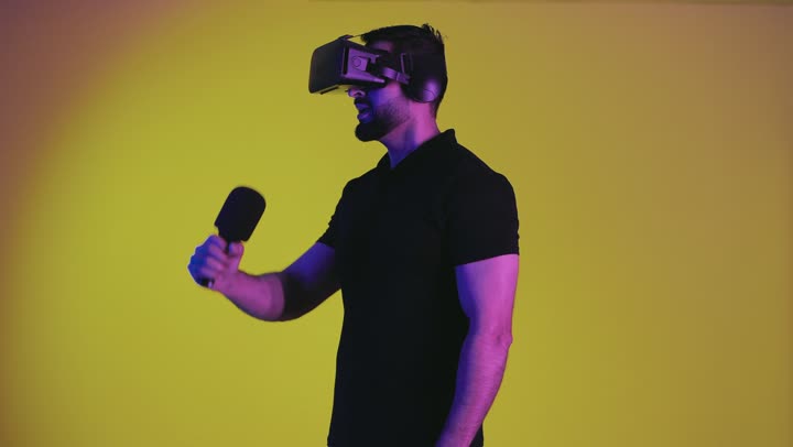 A young Saudi Arabian Gulf man wearing a 3D virtual reality headset holding a microphone and singing with gestures indicating happiness, entertainment and amusement by singing in the virtual world, talent and art, an Emirati man wearing casual clothes enjoying his singing, yellow background