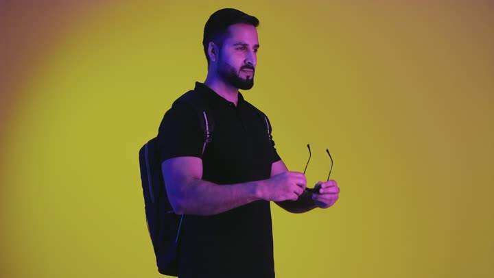 Portrait of a young Saudi Arabian Gulf man ready to travel wearing a backpack and sunglasses watching something on a sightseeing tour, Emirati man having a fun and entertaining trip, quality and excellence sign, convalescence and travel, yellow background