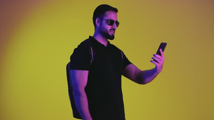 Portrait of a young Saudi Arabian Gulf man ready to travel wearing a backpack and sunglasses watching something on a sightseeing tour, Emirati man having a fun recreational trip, convalescence and travel, yellow background