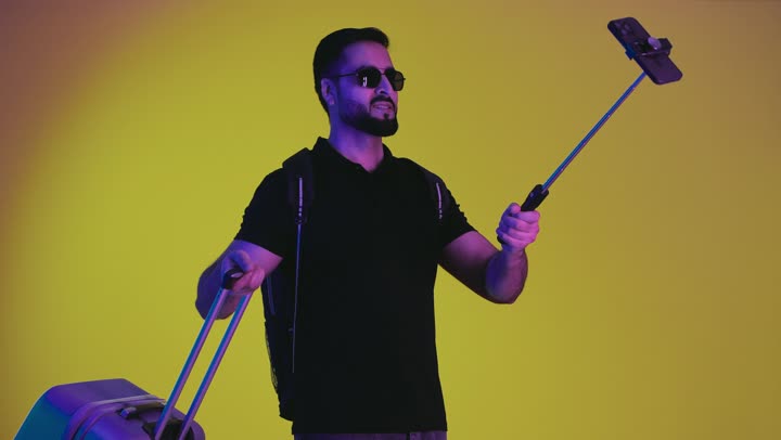 Having a fun and entertaining trip, portrait of a young Saudi Arabian Gulf man ready to travel holding a selfie stick, smiling Emirati man wearing sunglasses and carrying a suitcase taking souvenir photos during the tour, convalescence and travel, yellow background