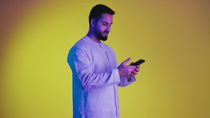 Using a mobile phone to communicate with people, making a phone call using a mobile phone, a picture of an Emirati Gulf Arab man wearing an Emirati kandura, chatting, conversations and getting to know each other, yellow background.