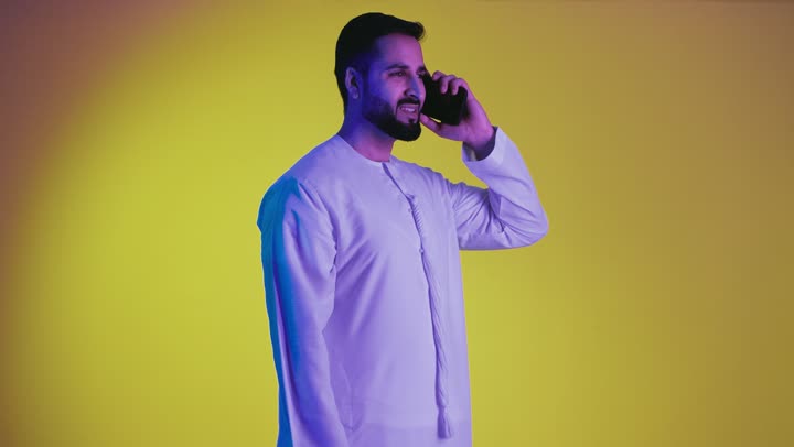 Making a phone call on a mobile phone, a picture of an Emirati Gulf Arab man wearing an Emirati kandura, using a mobile phone to call and communicate with individuals, chatting, conversations and getting to know each other, yellow background.
