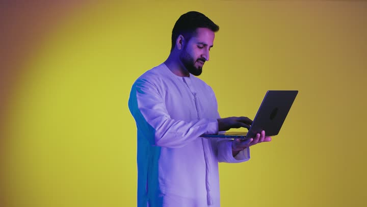 إدارة شؤون الأعمال وتنظيمها ، استخدام التقنيات الحديثة والأجهزة المتطورة ، مفهوم تسهيل الاعمال والقدرة على إنجاز المهام عن بعد، تصوير لشاب عربي خليجي اماراتى يرتدي الكندورة يمسك بيده حاسوب محمول تظهر عليه علامات الفرح والسرور، خلفية صفراء .
