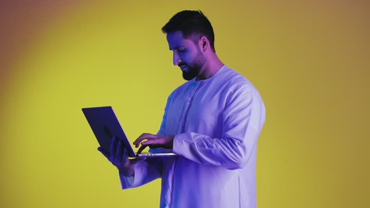 Remote work, use of modern technologies and advanced devices, the concept of facilitating work and the ability to complete tasks remotely, a picture of a young Emirati Gulf Arab man wearing a kandura holding a laptop, showing signs of joy and happiness, yellow background.