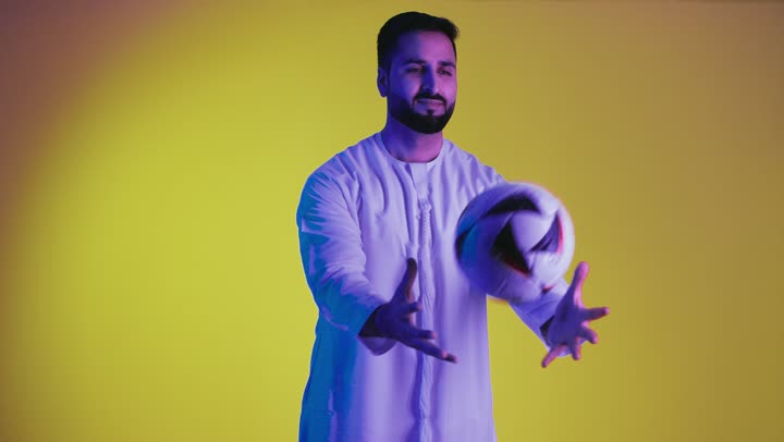 Facial and hand gestures indicating happiness, hobby of playing football, a picture of an Emirati Arab man from the Gulf wearing the Emirati kandura and holding a football in his hand, passion for football, clubs and cheering, yellow background.