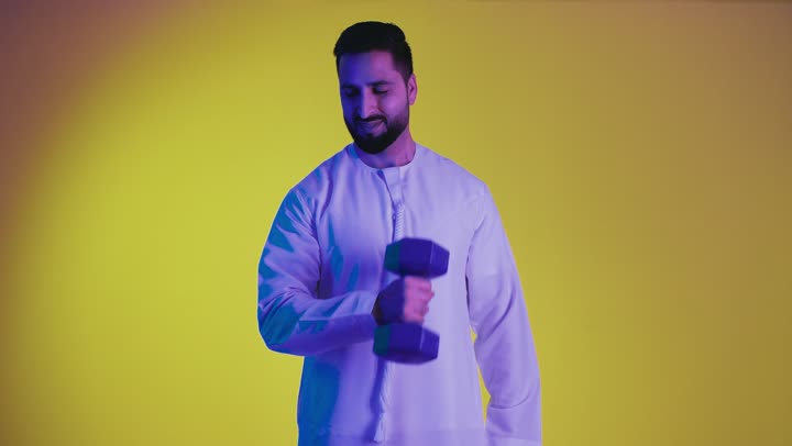 التمتع بالبنية الجسدية القوية، رفع الاوزان وعرض عضلات اليد ، تصوير لرجل عربي خليجي إماراتي يرتدي الكندورة الإماراتية ، إيماءات وجه ويدين تدل على القوة العضلية، اللياقة البدنية والجسم الصحي المتين، خلفية صفراء .