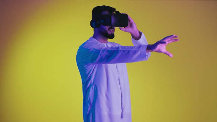 دمج العالم الخيالي ، تعابير الاندماج والتفاعل ، العالم الترفيهى الرقمى ، أجهزة تكنولوجية وتقنيات حديثة، تصوير لرجل عربي خليجي اماراتي مبتسم يرتدي الكندورة التقليدية ونظارة الواقع الإفتراضي يمسك بيده عصا التحكم يقوم باللعب في ألعاب الفيديو الترفيهية، خلفية صفراء .