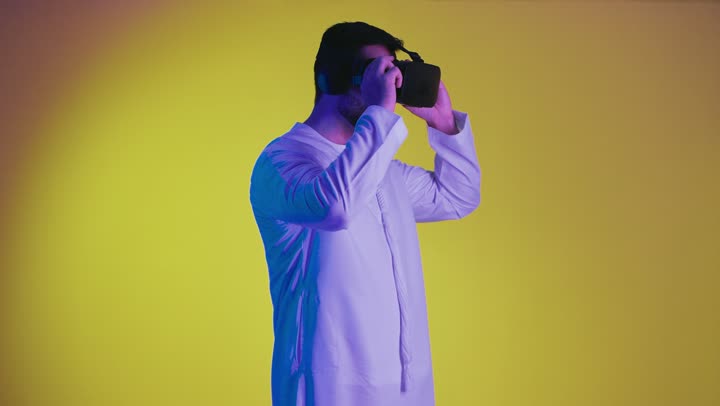 Expressions of integration and interaction, digital entertainment world, technological devices and modern techniques, integration of the imaginary world, a picture of a smiling Emirati Gulf Arab man wearing a traditional kandura and virtual reality glasses, holding a joystick in his hand, playing entertainment video games, yellow background.