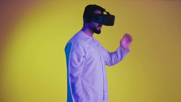 Fun and entertainment on vacation, digital entertainment world, technological devices and modern techniques, integration of the imaginary world, a picture of a smiling Emirati Gulf Arab man wearing the traditional kandura and virtual reality glasses holding a joystick in his hand playing entertainment video games, yellow background.