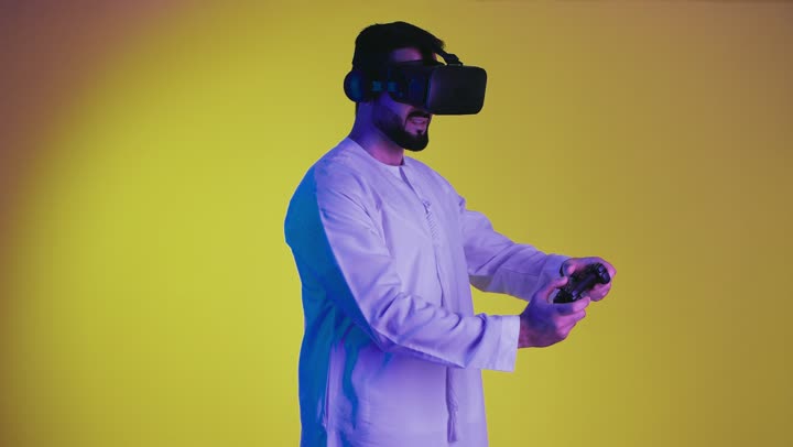 Digital entertainment world, technological devices and modern techniques, combining the imaginary world with fun and entertainment, a picture of a smiling Emirati Gulf Arab man wearing the traditional kandura and virtual reality glasses, holding a joystick in his hand, playing entertainment video games, yellow background.