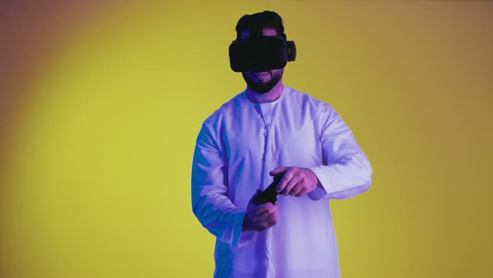 دمج العالم الخيالي مع المتعة والتسلية، تصوير لرجل عربي خليجي اماراتي مبتسم يرتدي الكندورة التقليدية ونظارة الواقع الإفتراضي يمسك بيده عصا التحكم يقوم باللعب في ألعاب الفيديو الترفيهية، أجهزة تكنولوجية وتقنيات حديثة، خلفية صفراء .