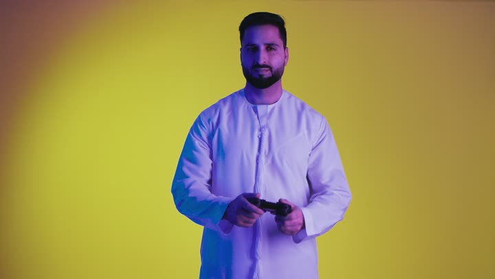 Having a good time, expressions of enjoyment while playing entertainment games, using technological devices and modern techniques, a picture of an Emirati Gulf Arab man wearing a kandura standing and holding a joystick with integration gestures, yellow background.
