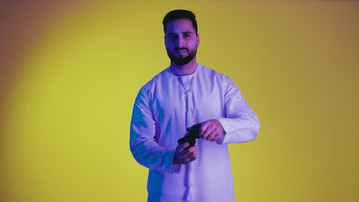 Expressions of enjoyment while playing recreational electronic games, using technological devices and modern techniques, a picture of an Arab Gulf Emirati man wearing a kandura standing and holding a joystick with integration gestures, yellow background.