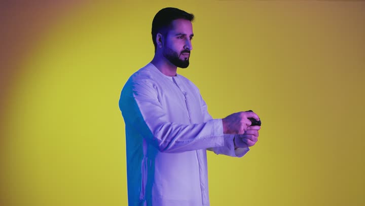 Using modern technological devices and techniques, expressions of enjoyment while playing recreational electronic games, a picture of an Emirati Gulf Arab man wearing a kandura and ghutra standing and holding a joystick with integration gestures, yellow background.